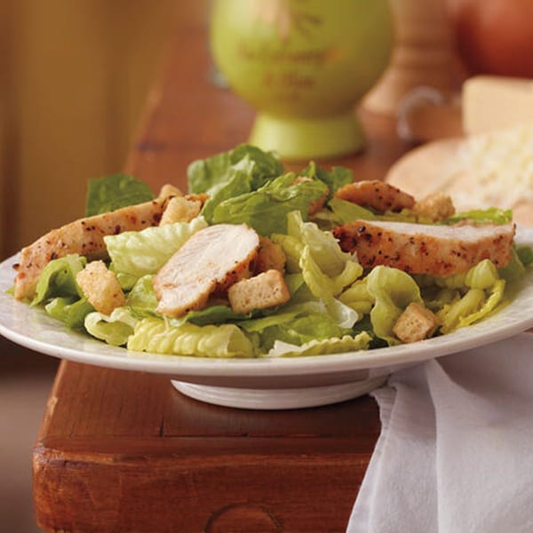 Chicken Caesar salad with croutons on a white plate, with a green pitcher and cheese in the background.